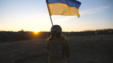Soldatin der ukrainischen Armee mit wehender Fahne der Ukraine. Frau in Militäruniform hisste Flagge gegen Sonnenuntergang. Sieg über die russische Aggression. Invasionswiderstandskonzept. Zeitlupe.