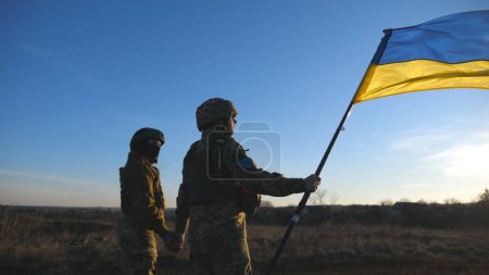 Ukrainian army soldiers stands outdoor at sunset time and waves flag of Ukraine. People in military uniform lifted up flag against blue sky. Victory at war. Resistance to russian invasion. Slow motion.
