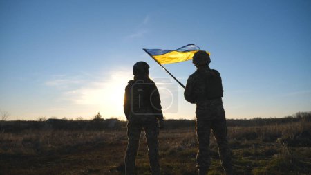 Ukrainian army soldiers stands outdoor at sunset time and waves flag of Ukraine. People in military uniform lifted up flag against blue sky. Victory at war. Resistance to russian invasion. Slow motion.