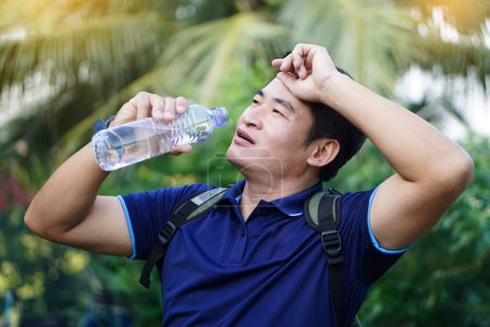 Foto de Hombre asiático guapo sostiene botella de agua potable para beber al aire libre, se siente caliente y cansado. Concepto: Agua potable para la salud, estilo de vida saludable.Apagando la sed, reducir la fatiga, refrescar el cuerpo. - Imagen libre de derechos