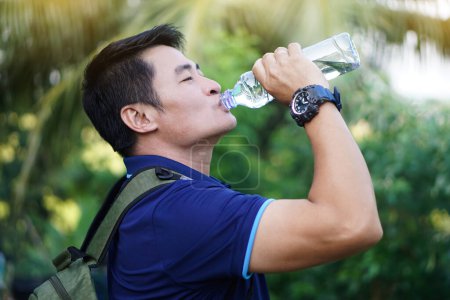 Foto de Guapo hombre asiático viajero sostiene botella de agua potable para beber al aire libre. Concepto: Agua potable para la salud, estilo de vida saludable.Apagando la sed, reducir la fatiga, refresca el cuerpo. - Imagen libre de derechos