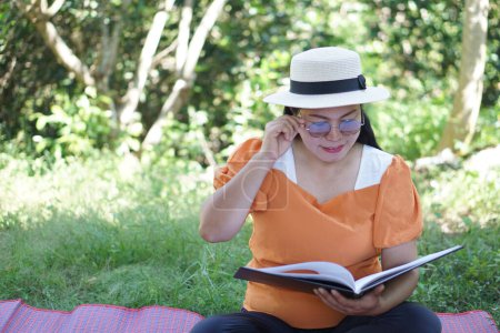 Photo for Asian middle- aged woman sits at the park, reads book, holds eyeglasses, has difficulties to see text because of bad vision. Concept , Eyesight problem. suffering eyestrain from reading  book. - Royalty Free Image