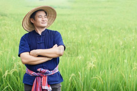 Photo for Handsome Asian man farmer is at paddy field, wears hat, blue shirt, Thai loincloth tied around waist, cross arms on chest.  Concept, agriculture occupation, Thai farmer. - Royalty Free Image