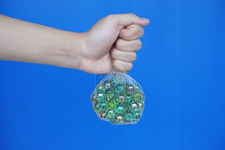 Aus nächster Nähe halten Sie Netztasche mit Murmelbällen, kleinen kugelförmigen Gegenständen, die oft aus Glas bestehen. Blauer Hintergrund. Konzept. Objektverwendung als Spielzeug oder zur Basteldekoration.