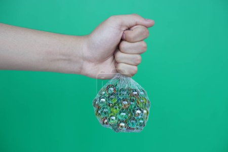 Aus nächster Nähe halten Sie Netztasche mit Murmelbällen, kleinen kugelförmigen Gegenständen, die oft aus Glas bestehen. Grüner Hintergrund. Konzept. Objektverwendung als Spielzeug oder zur Basteldekoration.