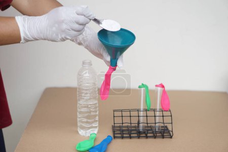 Closeup hands wears gloves holds funnel and flat pink balloon, put spoon of baking soda powder to pour into balloon. Concept, science experiment about reaction of chemical substance .  