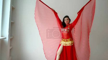 Eine Tänzerin im roten Anzug tanzt mit roten Flügeln einen orientalischen Tanz auf weißem Hintergrund. Freiraum für Text. Banner. Eine professionelle Tänzerin tanzt orientalischen Bauchtanz. Sexy Frau im roten Kleid