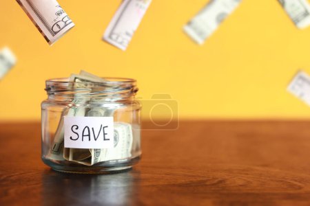 Photo for Save money for the future. Glass jar with dollars on a wooden table. Piggy bank with banknotes and dollars flying around. copy space. The concept of financial savings - Royalty Free Image