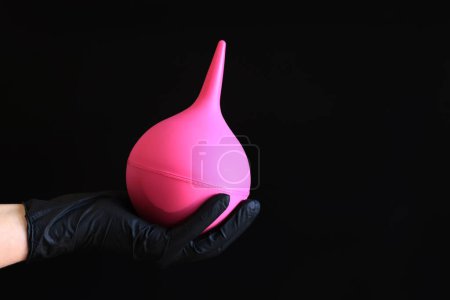Eine Hand in einem schwarzen Latex-Handschuh hält einen großen rosa Einlauf auf dunklem Hintergrund. Medizinprodukt. Birnenförmiges Einlauf-Konzept zur Körperreinigung