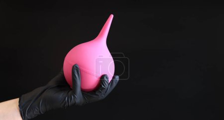 Eine Hand in einem schwarzen Latex-Handschuh hält einen großen rosa Einlauf auf dunklem Hintergrund. Medizinprodukt. Birnenförmiges Einlauf-Konzept zur Körperreinigung