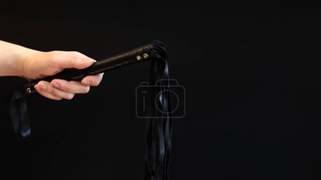 Photo for A woman's hand holds a whip against a dark background. Leather whip for spanking isolated on black. Sex toy for intimacy. Sexual slavery - Royalty Free Image