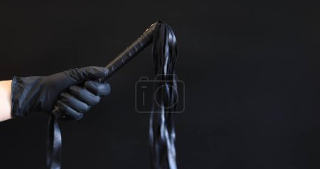 Photo for A hand in a black latex glove holds a whip against a dark background. Leather whip for spanking isolated on black. Sex toy for intimacy. Sexual slavery - Royalty Free Image