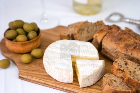 Photo for Camembert cheese, sliced fresh baguette, olives on a wooden board on the table. A wineglass and a bottle of white wine blurred in the background. Close-up. Selective focus. - Royalty Free Image