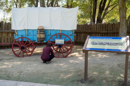 Foto de Génova, NV, EE.UU. 2022-09-17. Joven no identificado mirando un vagón azul Prairie Schooner con ruedas rojas en el Parque Histórico de la Estación Mormón. - Imagen libre de derechos