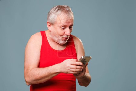 Foto de Senior Citizen Technology Shock A senior man in a studio environment is captured gazing at his smartphone, as if he is in a state of shock. He appears to be flabbergasted and bewildered - Imagen libre de derechos