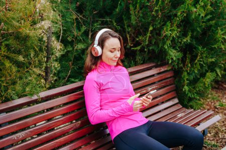 Photo for A pretty caucasian woman sits in a chair in the park, surrounded by lush greenery, and takes a moment to relax. With her smartphone nearby, enjoy her favorite tunes through her headphones - Royalty Free Image