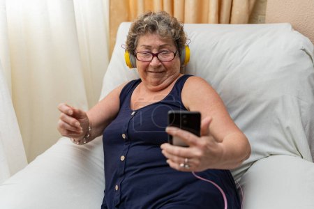Téléchargez les photos : Femme âgée avec écouteurs regardant smartphone dans le salon. Concept de personnes âgées utilisant la technologie. - en image libre de droit