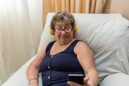 Téléchargez les photos : Femme plus âgée avec écouteurs regardant son téléphone intelligent tout en étant assis sur le canapé à la maison. Concept de personnes âgées utilisant la technologie. - en image libre de droit