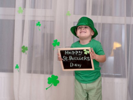 Photo for St. Patricks Day concept. A small cheerful caucasian boy in a shiny green top hat and green clothes holds a black wooden board with an inscription in chalk the holiday of St. Patrick on the - Royalty Free Image