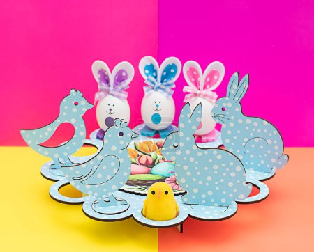 Téléchargez les photos : Concept de Pâques. Stand festif pour les œufs de Pâques. Œufs décoratifs avec des muselières drôles peintes et des oreilles en tissu. Figurines en bois sous la forme d'un poulet et un lapin de Pâques sur un lumineux multicolore - en image libre de droit