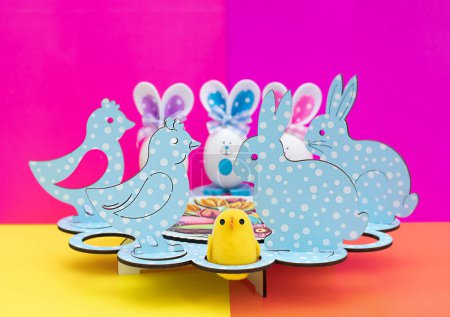 Téléchargez les photos : Concept de Pâques. Stand festif pour les œufs de Pâques. Œufs décoratifs avec des muselières drôles peintes et des oreilles en tissu. Figurines en bois sous la forme d'un poulet et un lapin de Pâques sur un fond multicolore lumineux. - en image libre de droit