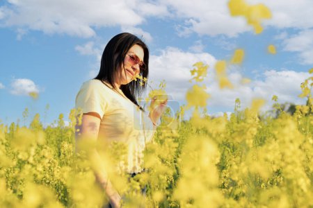 Téléchargez les photos : Une belle fille en lunettes de soleil se tient dans un champ de fleurs jaunes contre un ciel bleu, sourit et sent une fleur de colza. Concept de paix. Concentrez-vous. espace de copie. - en image libre de droit