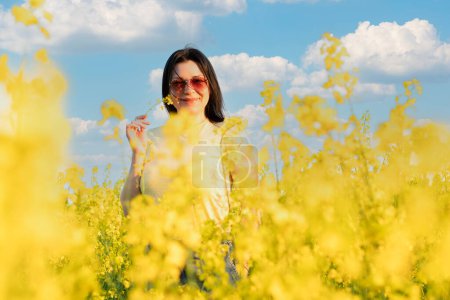 Téléchargez les photos : Une belle fille en lunettes de soleil se tient dans un champ de fleurs jaunes contre un ciel bleu, sourit joyeusement et respire le printemps frais et l'air pur. Concept de paix. La couleur du drapeau de l'Ukraine. Non. - en image libre de droit