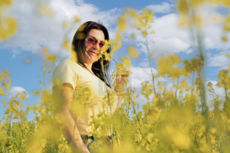 Téléchargez les photos : Une belle fille en lunettes de soleil se tient dans un champ de fleurs jaunes contre un ciel bleu, sourit joyeusement et respire le printemps frais et l'air pur. Concept de paix. La couleur du drapeau de l'Ukraine. Non. - en image libre de droit