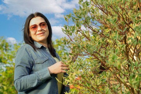 Photo for Camping. A beautiful girl in jeans and sunglasses, stands near a blooming green tree, smiles happily. Fresh air concept. Sunset. - Royalty Free Image