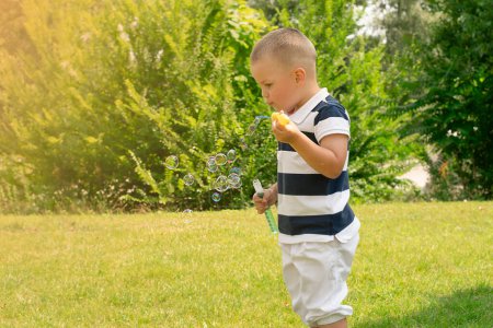 Téléchargez les photos : Un beau garçon caucasoïde de 4 ans, en t-shirt rayé, joue à l'extérieur dans un parc verdoyant en été et souffle des bulles de savon au soleil. Concept d'enfance. - en image libre de droit