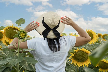 Téléchargez les photos : Un symbole de paix. Concept Ukraine. Une fille dans un chapeau de paille se tient dans un champ avec des tournesols jaunes contre un ciel bleu. Vue de l'arrière. Pas de visage. Gros plan. - en image libre de droit