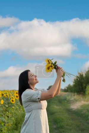 Téléchargez les photos : Concept Ukraine. Un symbole de paix. Une belle et heureuse fille caucasienne se tient dans un champ de tournesols jaunes contre un ciel bleu et tient un bouquet de fleurs dans ses mains. - en image libre de droit