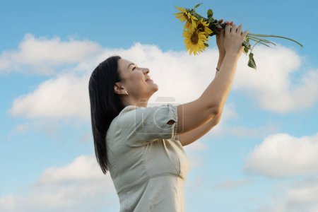Téléchargez les photos : Un symbole de paix. Une belle et heureuse fille caucasienne se tient contre le ciel bleu et tient un bouquet de fleurs avec des tournesols jaunes dans ses mains. Ukraine concept. - en image libre de droit