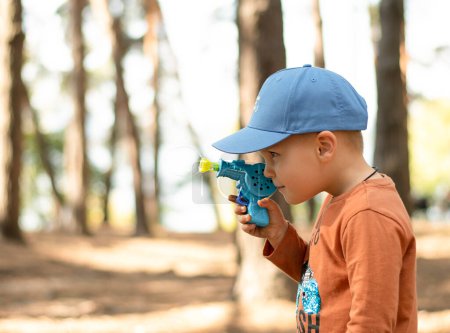 Téléchargez les photos : Enfants dans la nature. Un petit garçon beau et émotionnel dans une casquette bleue et une veste rouge joue avec des bulles de savon dans l'air frais de la forêt. - en image libre de droit