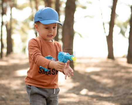 Téléchargez les photos : Enfants dans la nature. Un petit garçon beau et émotionnel dans une casquette bleue et une veste rouge joue avec des bulles de savon dans l'air frais de la forêt. - en image libre de droit