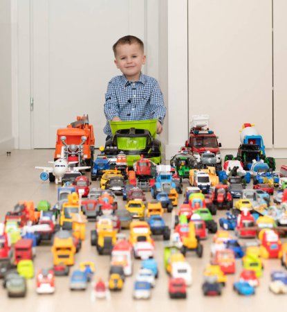 Photo for Concept of childrens toys. A little boy, 4 years old, cheerful and handsome, happily plays, sitting on the floor, with multi-colored small and large cars in the childrens room. Soft focus - Royalty Free Image
