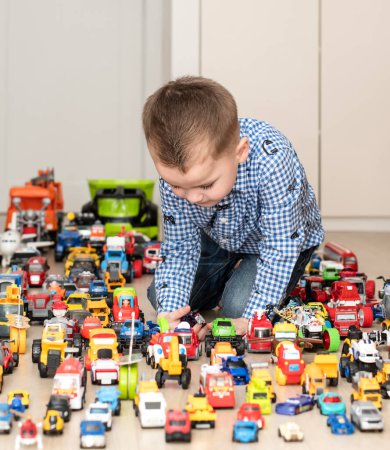 Photo for Concept of childrens toys. A little boy, 4 years old, enthusiastically plays, sitting on the floor, with multi-colored small and large cars in the childrens room. Soft focus - Royalty Free Image
