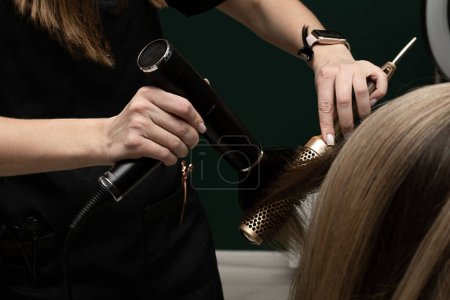 Beauty sphere. The master hairdresser does styling and combing the hair. Combs a client's long hair with a round brush and blow-dries it in a beauty salon. Close-up. Business concept. No faces.