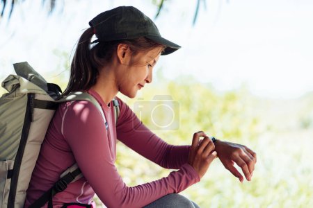 Photo for Solo Traveler backpacker sitting under the tree while take a break and checking smartwatch. Traveling outdoor in summer vacation. - Royalty Free Image