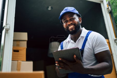 Foto de African courier man house mover Comprobación de caja de cartón con tableta antes de pasar al cliente. Hombre optimista auténtico feliz y disfruta de su trabajo. - Imagen libre de derechos