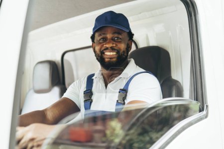 Foto de Retrato del mensajero negro en el camión sonriendo a la cámara mientras está sentado en el asiento del conductor, trabajador optimista con ocupación de entrega. - Imagen libre de derechos