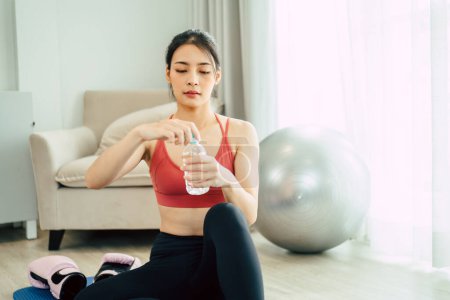 Foto de Mujer asiática en yoga ropa deportiva sentada en la esterilla y agua potable abierta después de entrenar yoga. - Imagen libre de derechos