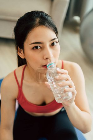 Foto de Mujer asiática en ropa deportiva de yoga sentada en la esterilla y bebiendo agua. - Imagen libre de derechos