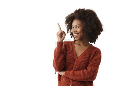 Foto de Mujer negra joven en suéter de punto rojo casual sonriendo señala con el dedo la pantalla izquierda que tiene espacio para copiar en el fondo blanco de aislamiento. la chica sonrisa y mirando a la cámara. - Imagen libre de derechos