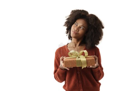 Téléchargez les photos : Jolie jeune femme afro-américaine en pull tricoté rouge tenant boîte cadeau sur fond mural blanc. Fille souriante, elle est heureuse d'obtenir un cadeau pour le festival de Noël et bonne année. - en image libre de droit