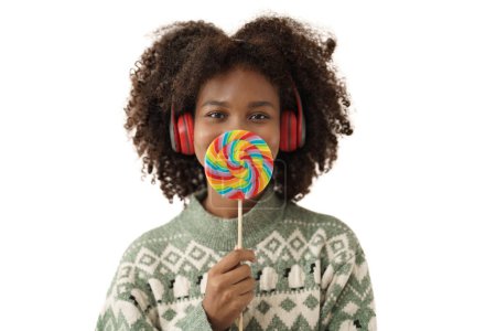 Téléchargez les photos : Mignonne afro-américaine jeune femme portant un pull tricoté tenant sucette devant sa bouche et souriant. - en image libre de droit