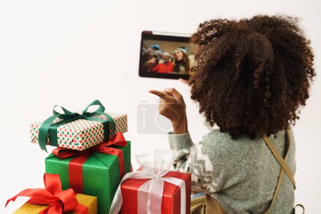 Foto de Mujer joven afroamericana en jersey de punto sosteniendo la tableta mientras llama en línea con su familia para mostrar la caja de regalo cerca, ella está feliz de recibir regalo para el festival de Navidad y feliz año nuevo. - Imagen libre de derechos