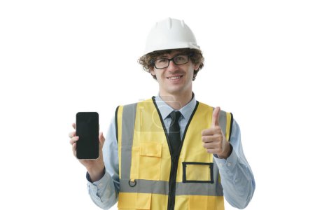 Foto de Retrato de un joven trabajador industrial, ingeniero con sombrero de señora sonriendo mostrando el teléfono con el pulgar hacia arriba, mirando feliz y seguro de sí mismo, ocupación profesional. - Imagen libre de derechos