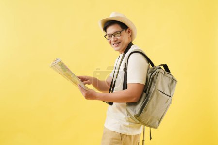 Téléchargez les photos : Un jeune voyageur asiatique portant un chapeau debout tout en tenant une carte et un sac de voyage avec des bagages, il regarde l'appareil photo et sourit. Vacances d'été et aventure concept de voyage. - en image libre de droit