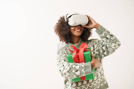 Foto de Joven mujer afroamericana con AR o gafas de realidad aumentada sonriente abrazo presente caja y mirar a la pantalla derecha con emocionante con el mundo virtual. - Imagen libre de derechos